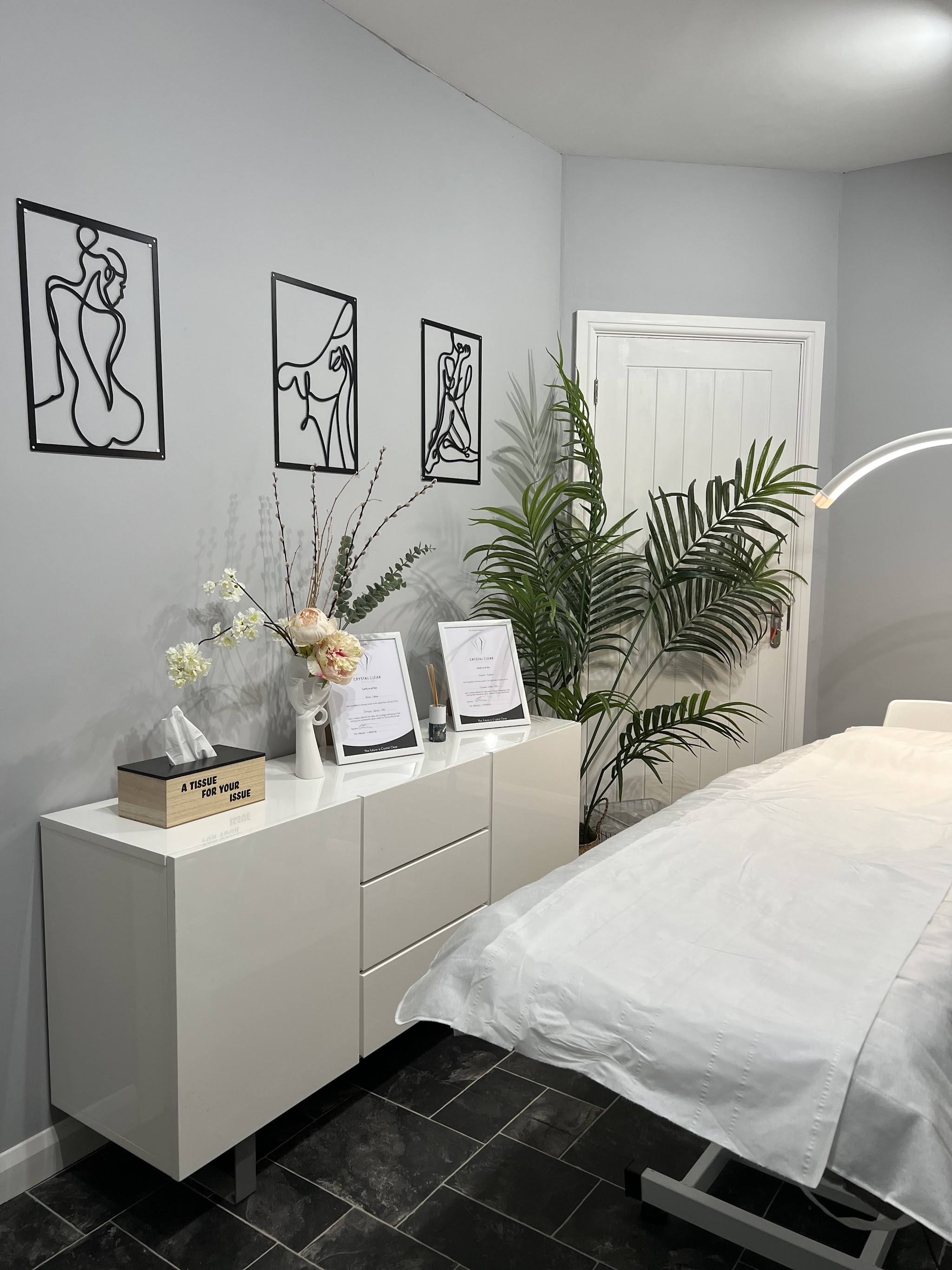 A treatment room with a white bed, white drawers and certificates on the side
