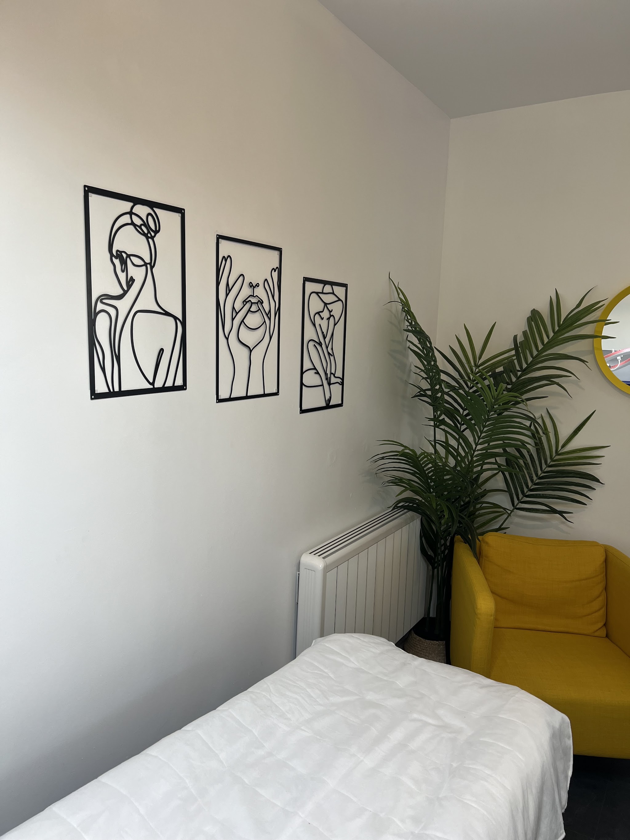 A light treatment room with a made-up treatment bed, a yellow armchair and three line-art prints